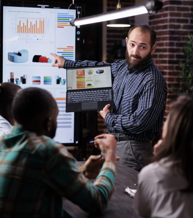 Startup entrepreneur holding laptop with sales presentation charts pointing at wall screen tv in late night meeting. Caucasian man presenting marketing strategy to coworkers working overtime.